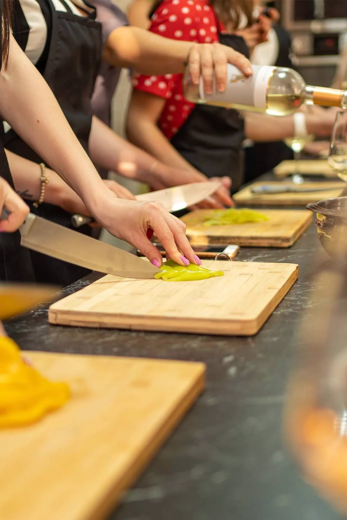 Hands slicing vegetables on wooden boards at a long worktop, with wine poured into a glass alongside.