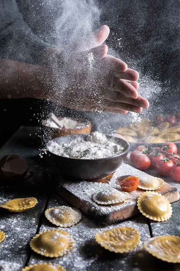 Floured hands above a bowl of flour, with fresh ravioli on a wooden board.